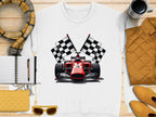 A Garment Graphics T-shirt, featuring a red racing car and two checkered flags on a white backdrop, is placed on a wooden surface. Perfect for motorsport fans, its accompanied by a scarf, notebook, pen, yellow wallet, sunglasses, yellow jacket, and coffee mug.