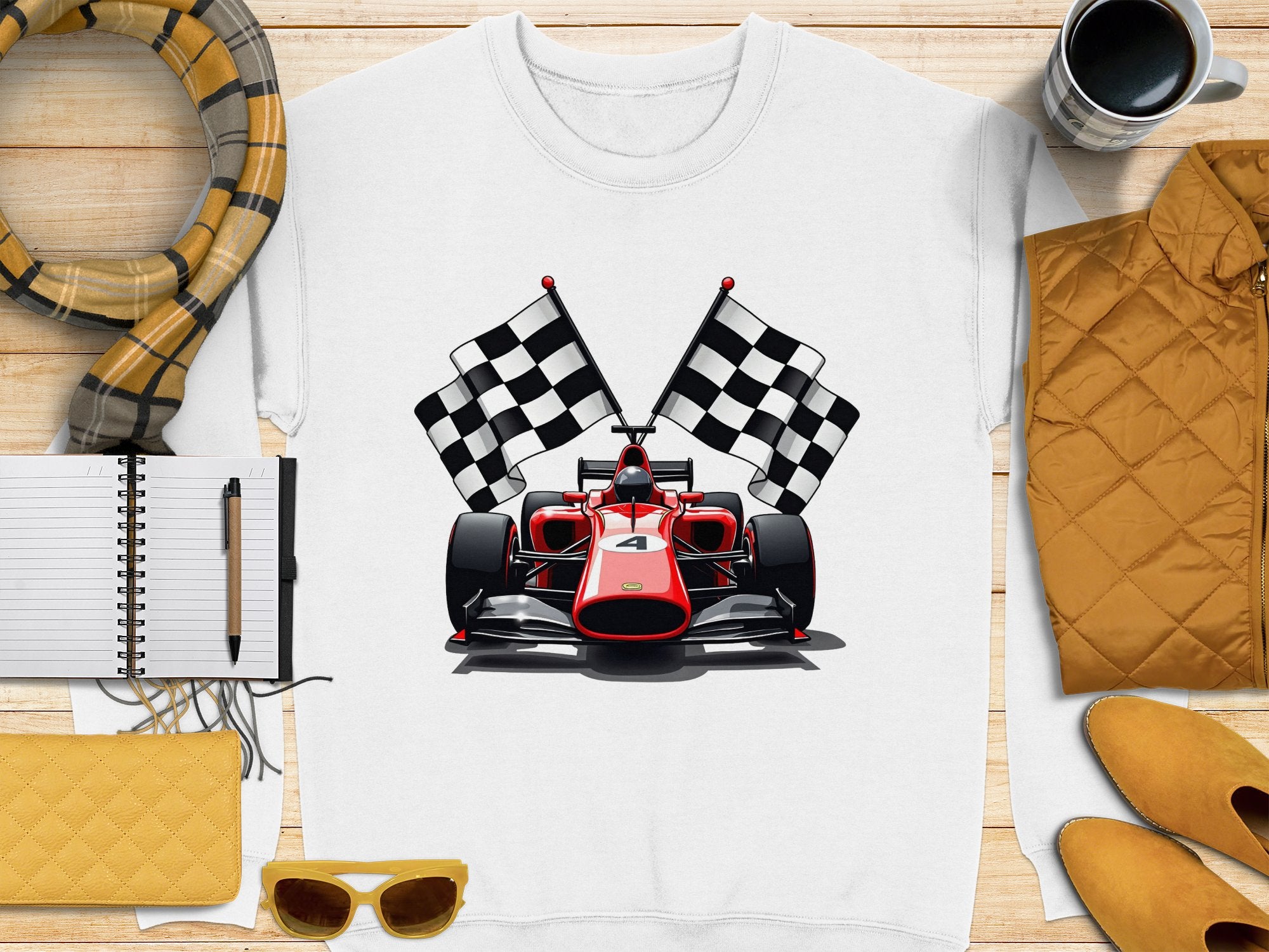 A Garment Graphics T-shirt, featuring a red racing car and two checkered flags on a white backdrop, is placed on a wooden surface. Perfect for motorsport fans, its accompanied by a scarf, notebook, pen, yellow wallet, sunglasses, yellow jacket, and coffee mug.