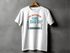Garment Graphics white fishing T-shirt features Id rather have a bad day fishing than a good day at work in orange and teal fonts. Ideal for anglers, it hangs on a wooden hanger against a gray background, making it an excellent gift for any fishing enthusiast.