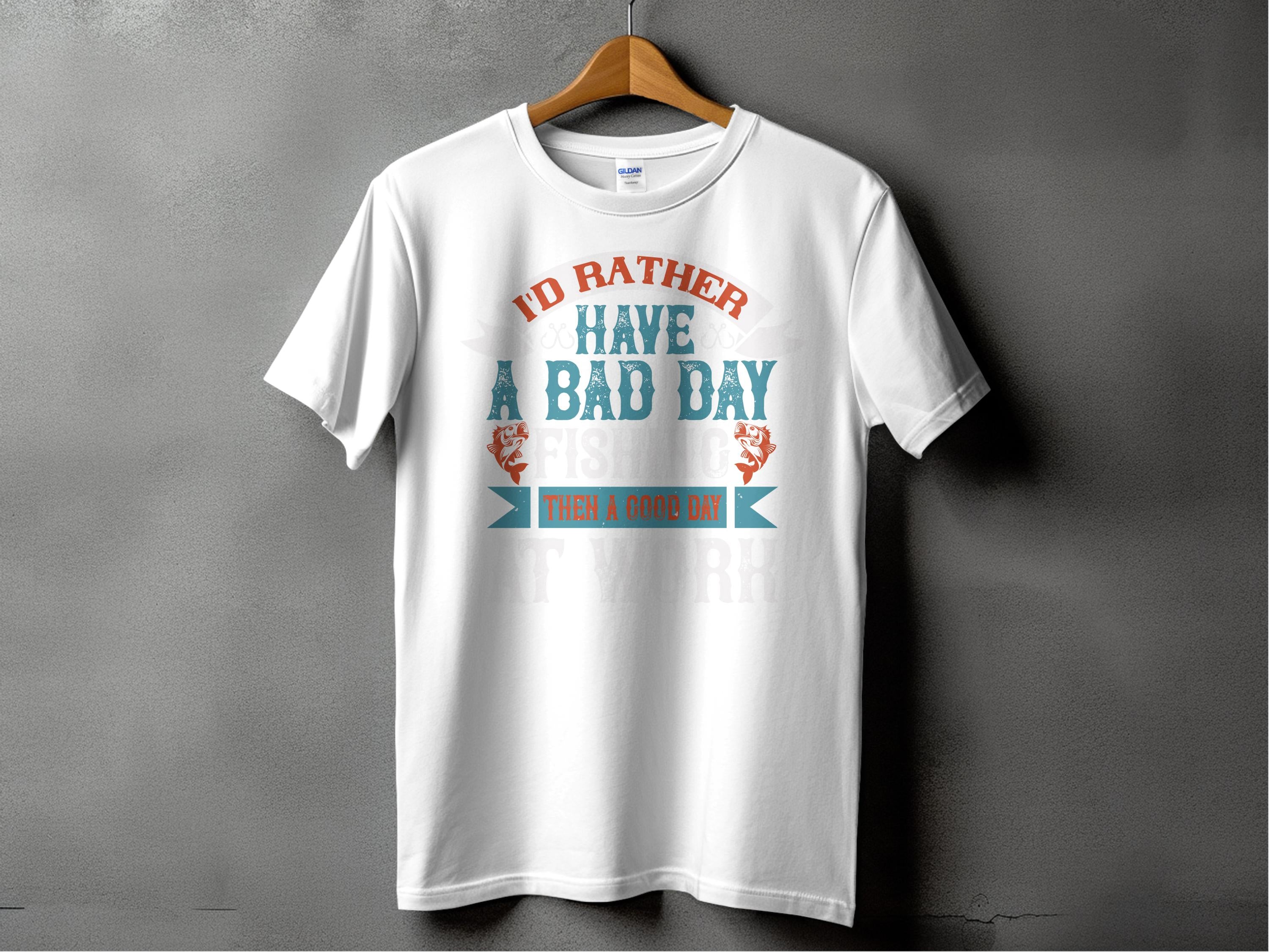 Garment Graphics white fishing T-shirt features Id rather have a bad day fishing than a good day at work in orange and teal fonts. Ideal for anglers, it hangs on a wooden hanger against a gray background, making it an excellent gift for any fishing enthusiast.