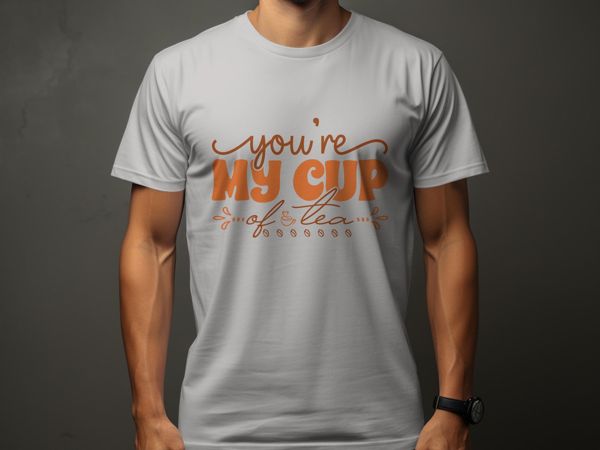 A person is wearing a light gray Garment Graphics T-shirt made of 100% cotton, featuring a playful Youre My Cup of Tea text in decorative brown lettering. The dark background highlights their relaxed pose.