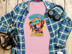 A Garment Graphics pink t-shirt features a vibrant dog-in-sunglasses graphic with the phrase Chillin Like A Canine Villain. Its creatively displayed on a wooden surface with a blue plaid shirt, black headphones, and a camera.