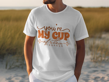 A person in a Garment Graphics 100% cotton T-shirt with Youre My Cup of Tea in stylish brown text stands outdoors against a blurred grass and sandy backdrop.