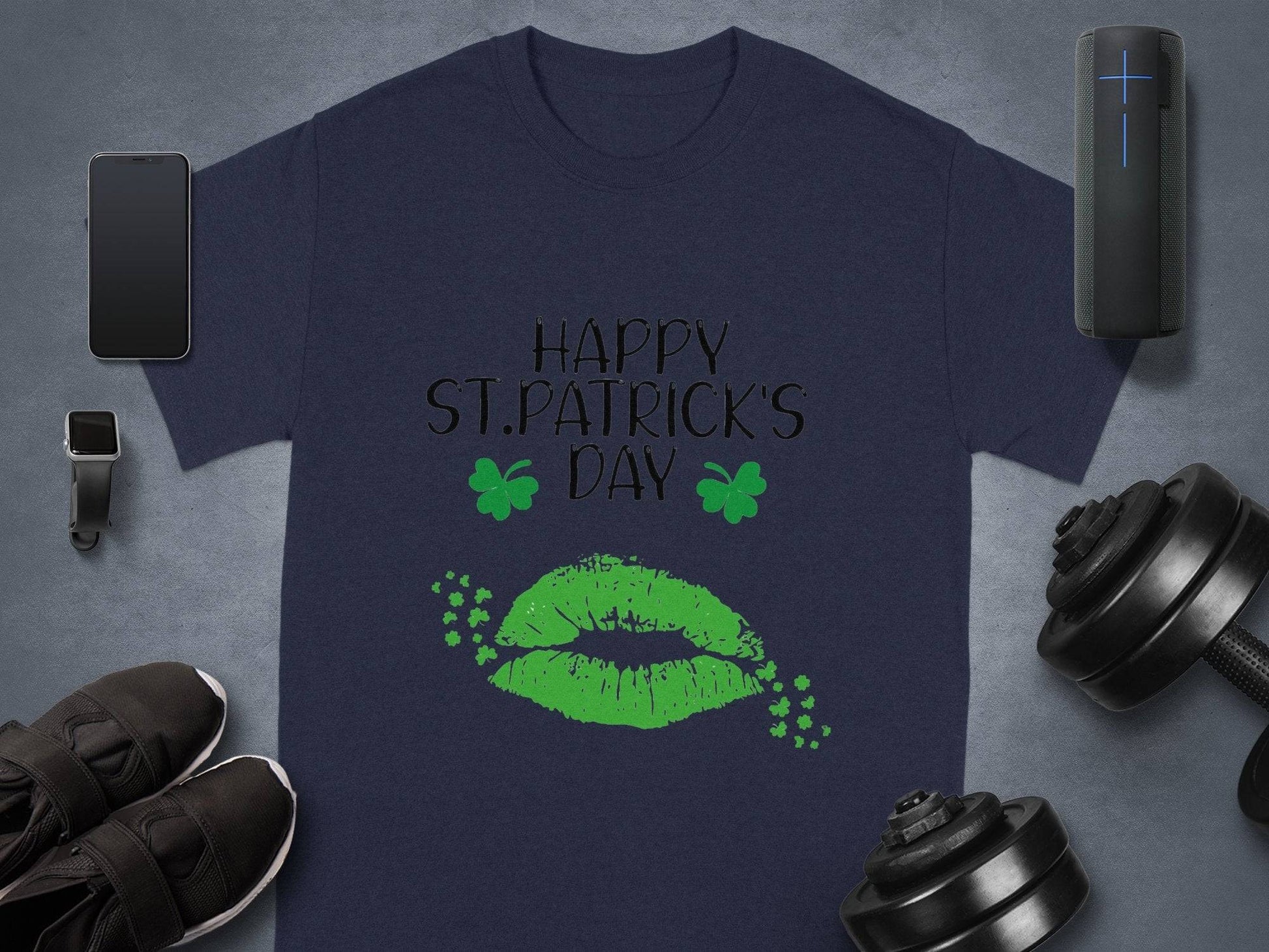 A navy blue Garment Graphics T-shirt showcases Happy St. Patricks Day with green lips and clovers. Nearby are a smartphone, smartwatch, wireless speaker, black shoes, and adjustable dumbbells.