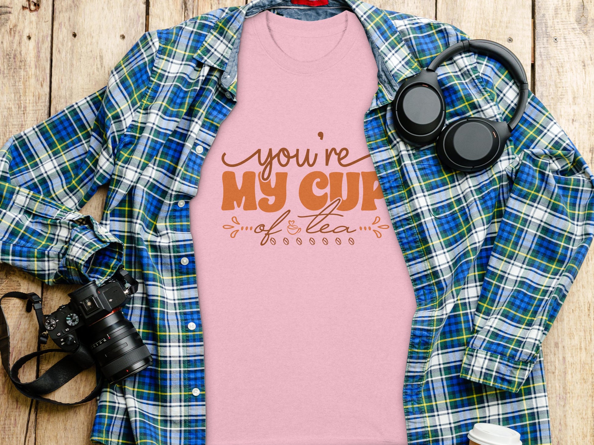 A flat lay showcases a Garment Graphics 100% cotton pink T-shirt with Youre My Cup of Tea in brown font, paired under an open blue and green plaid shirt. Nearby, headphones, a camera, and part of a white cup complete the scene.