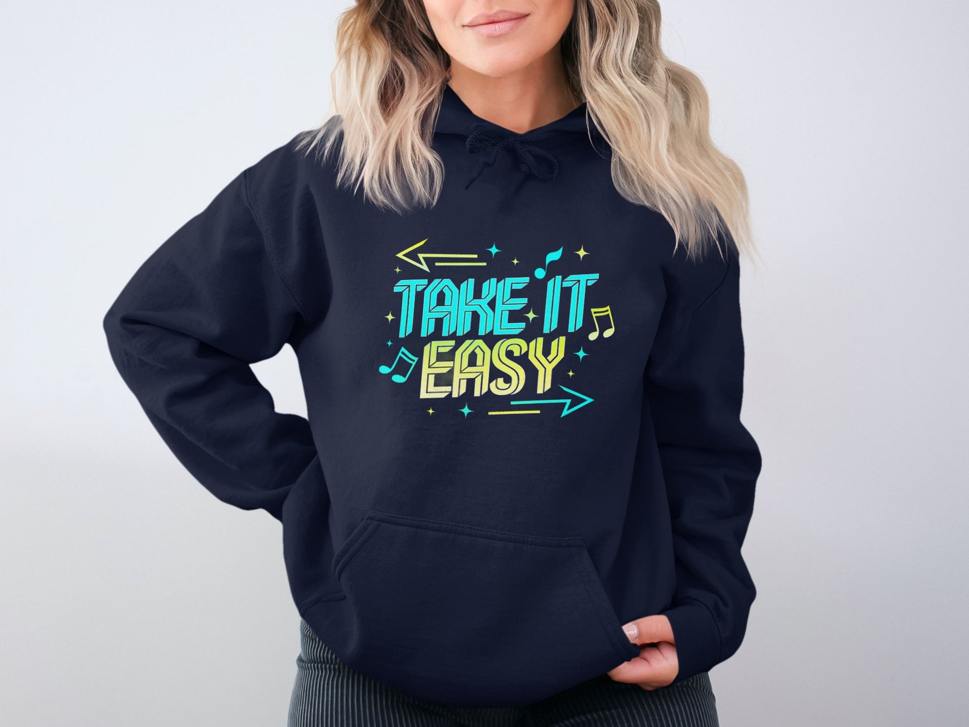 A wavy-haired blonde sports a Garment Graphics navy hoodie with Take it Easy in bright colors. Featuring musical notes and arrows, this comfy style exudes charm and ease as they relax with hands in the pockets.