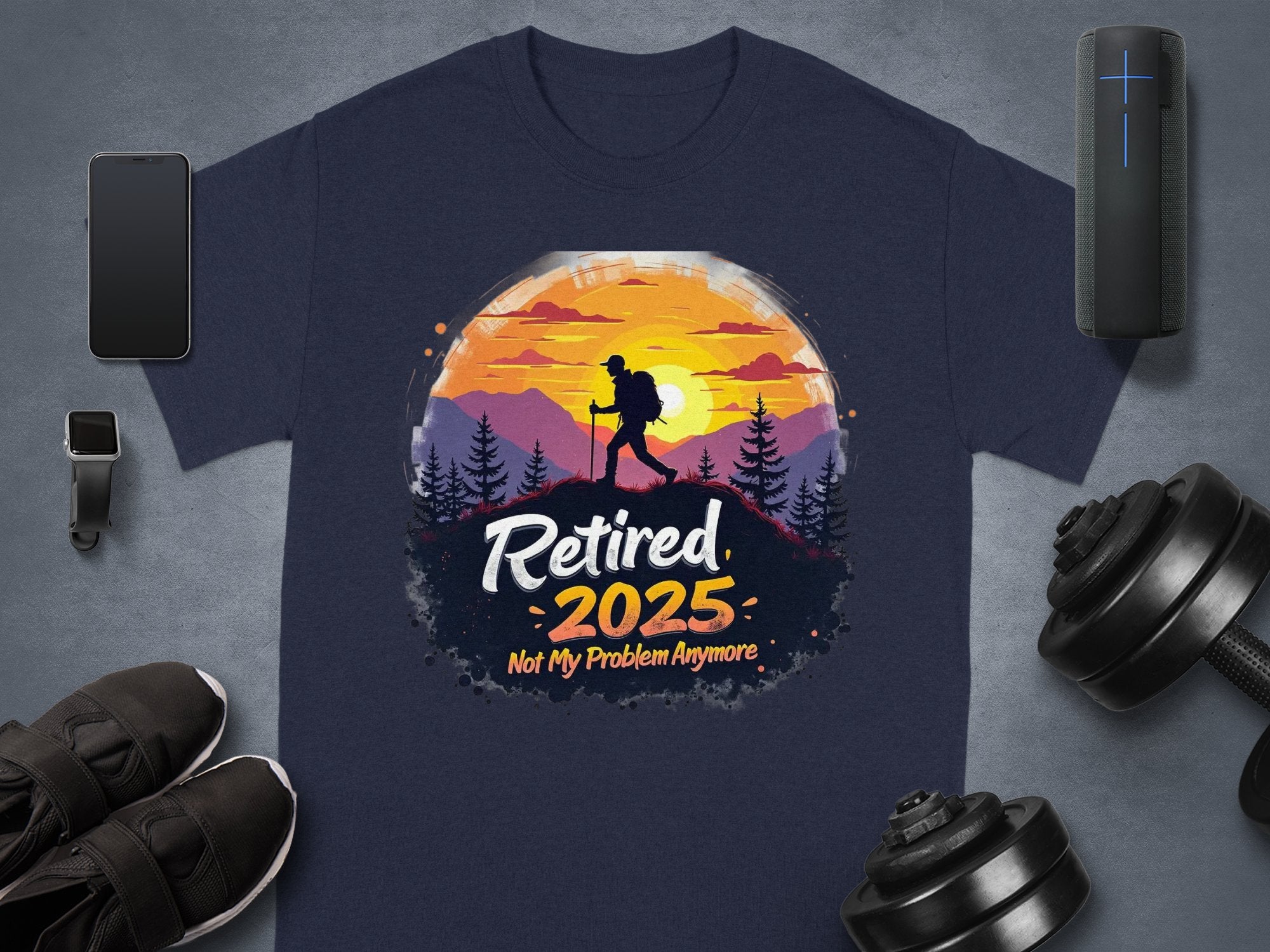 A Garment Graphics navy t-shirt features a hiker silhouette against a sunset with mountains and trees, celebrating Retired 2025—Not My Problem Anymore, perfect for relaxation, alongside essentials like a phone, smartwatch, shoes, weights, and speaker.