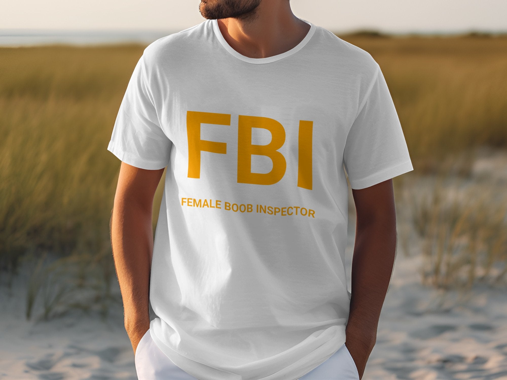 A person outdoors in a white Garment Graphics T-shirt, featuring FBI in yellow and Female Boob Inspector below, stands against a grassy landscape under an overcast sky, making for a funny tee that brings humor to any cloudy day.