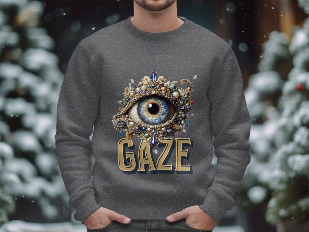 Someone is donning a gray sweatshirt by Garment Graphics in unisex sizing, featuring an intricate gem and ornament eye design above GAZE, set against a backdrop of snow-covered trees with gently falling snow.
