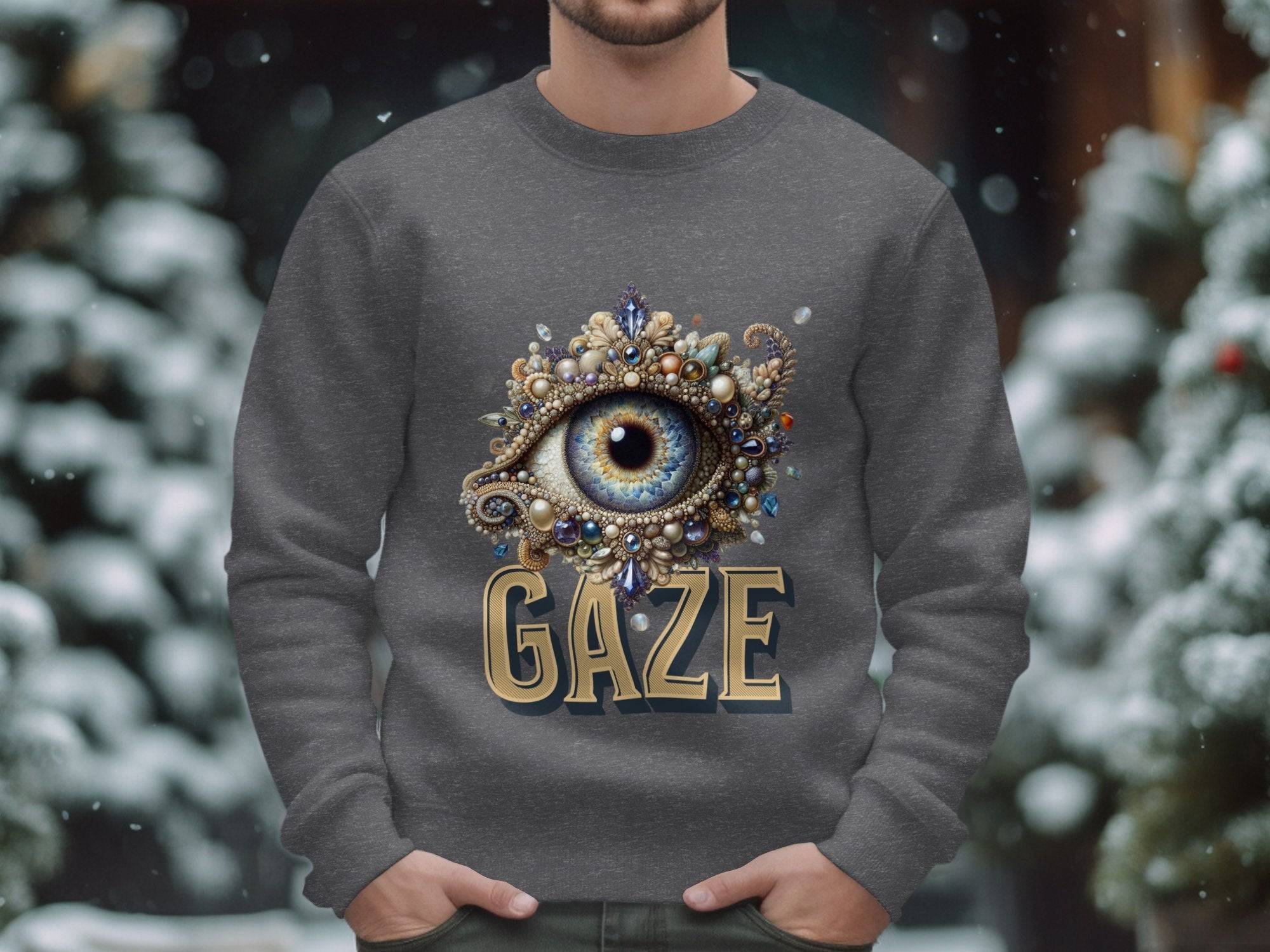 Someone is donning a gray sweatshirt by Garment Graphics in unisex sizing, featuring an intricate gem and ornament eye design above GAZE, set against a backdrop of snow-covered trees with gently falling snow.