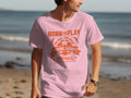 A man wearing a light pink football T-shirt by Garment Graphics with an orange soccer design stands on a beach. The shirt reads, Born to Play, One Goal, One Dream, Young Squad Football, with the ocean stretching in the background.