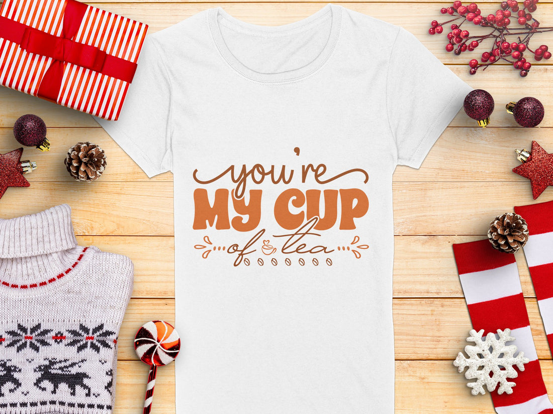 A woman wears a Garment Graphics 100% cotton graphic T-shirt featuring youre my cup of tea in brown. She stands outside against a backdrop of blurred trees and snow, with her hands cozily tucked into her pockets.