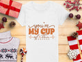 A flat lay showcases a Garment Graphics 100% cotton graphic T-shirt with youre my cup of tea in brown text, encircled by festive Christmas decor: red-wrapped gifts, pinecones, candies, a knitted sweater, and red stars on a wooden surface.