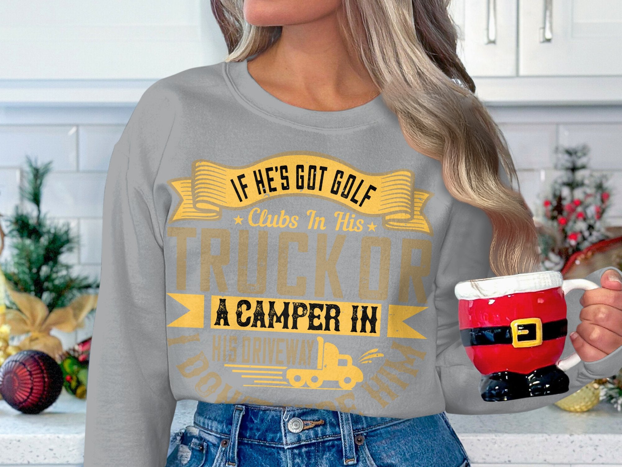 A person with long hair wears a Garment Graphics sweatshirt that humorously states, If hes got golf clubs in his truck or a camper in his driveway, I dont want him, and holds a festive Santa suit-shaped mug. Holiday decorations create a cozy atmosphere.