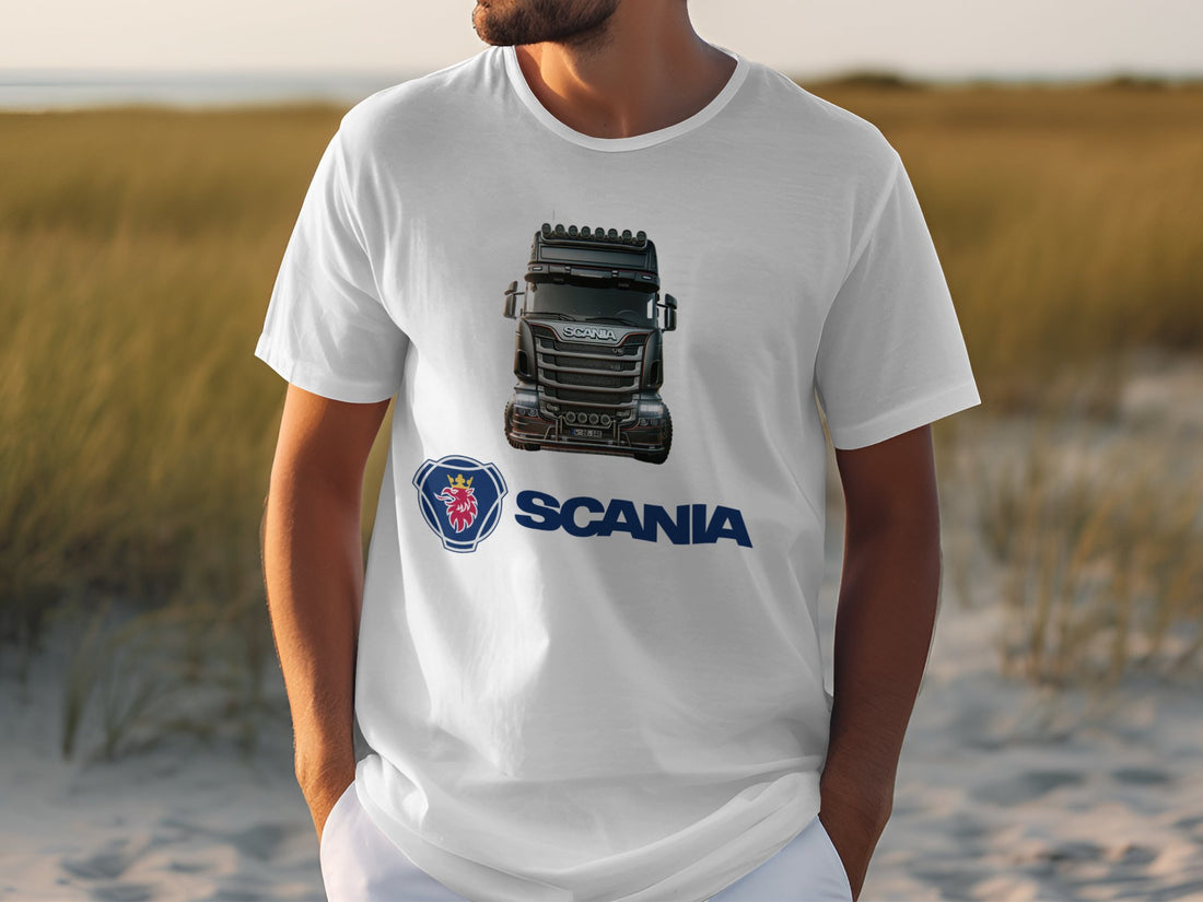 A person wears a Garment Graphics white T-shirt with a black Scania truck, ideal for truck lovers. The iconic Scania logo and name are beneath the image on a grassy field background.