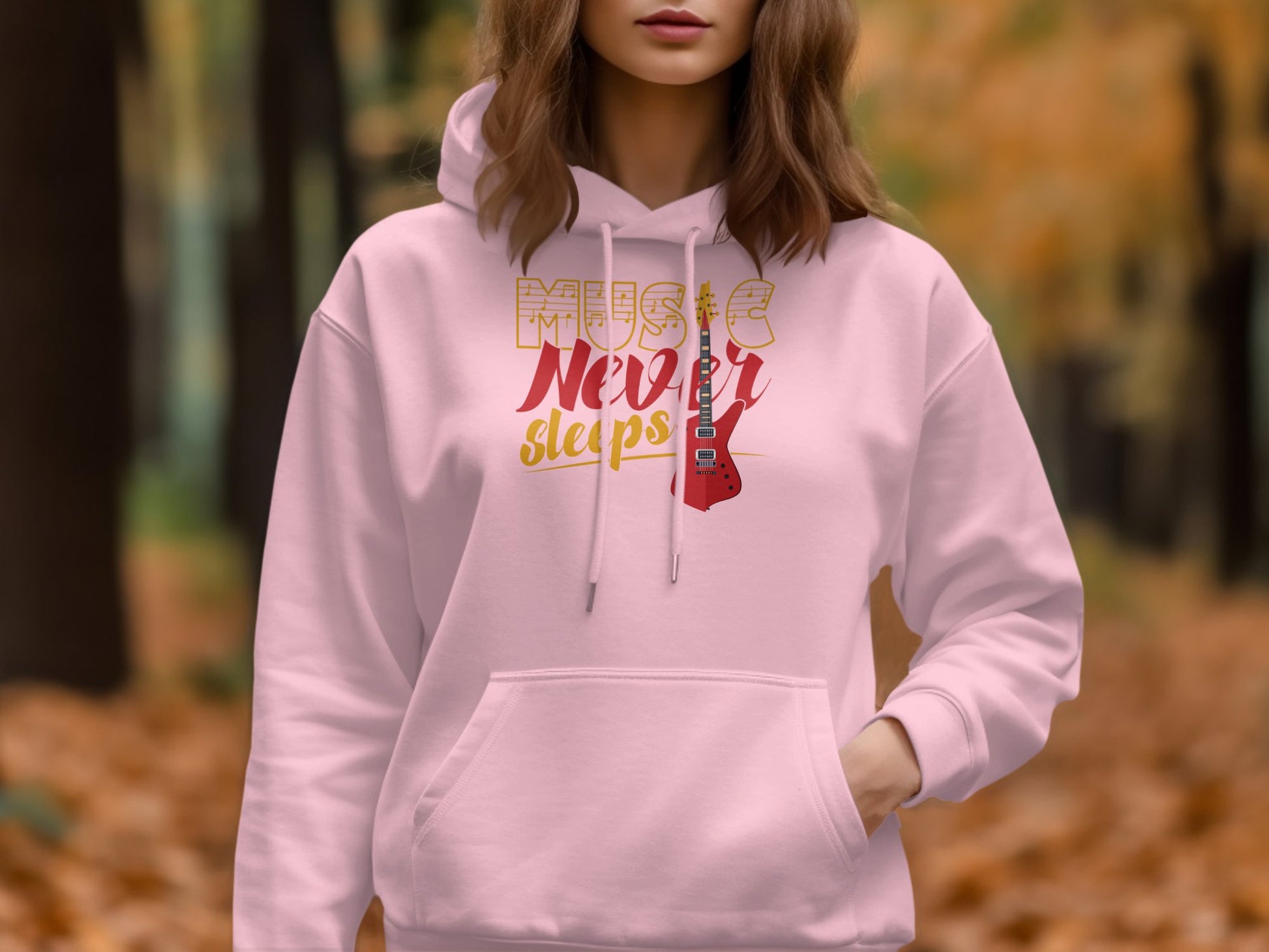 Someone is wearing a light pink, classic fit hoodie from Garment Graphics. The hoodie features the text Music Never Sleeps and a graphic guitar design, as they stand outdoors with autumn leaves in the background.