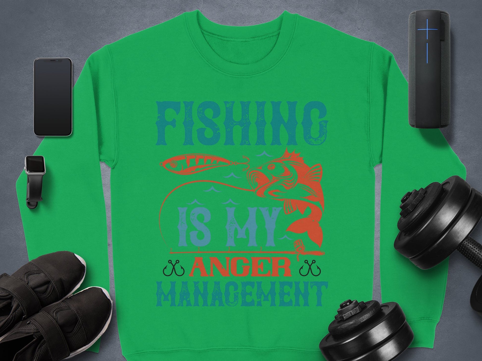 Garment Graphics offers a green sweatshirt for fishing fans, displaying Fishing Is My Anger Management with a fish graphic. Accompanied by items like a smartphone, earbuds, black sneakers, a portable speaker, and adjustable dumbbells on gray—ideal for casual wear.