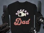 The Soccer Dad sweatshirt by Garment Graphics, showcasing a white and pink soccer ball design, pairs seamlessly with items like smartphones and black sneakers on a grey backdrop. This stylish, cozy piece offers unisex sizing for maximum comfort and versatility.
