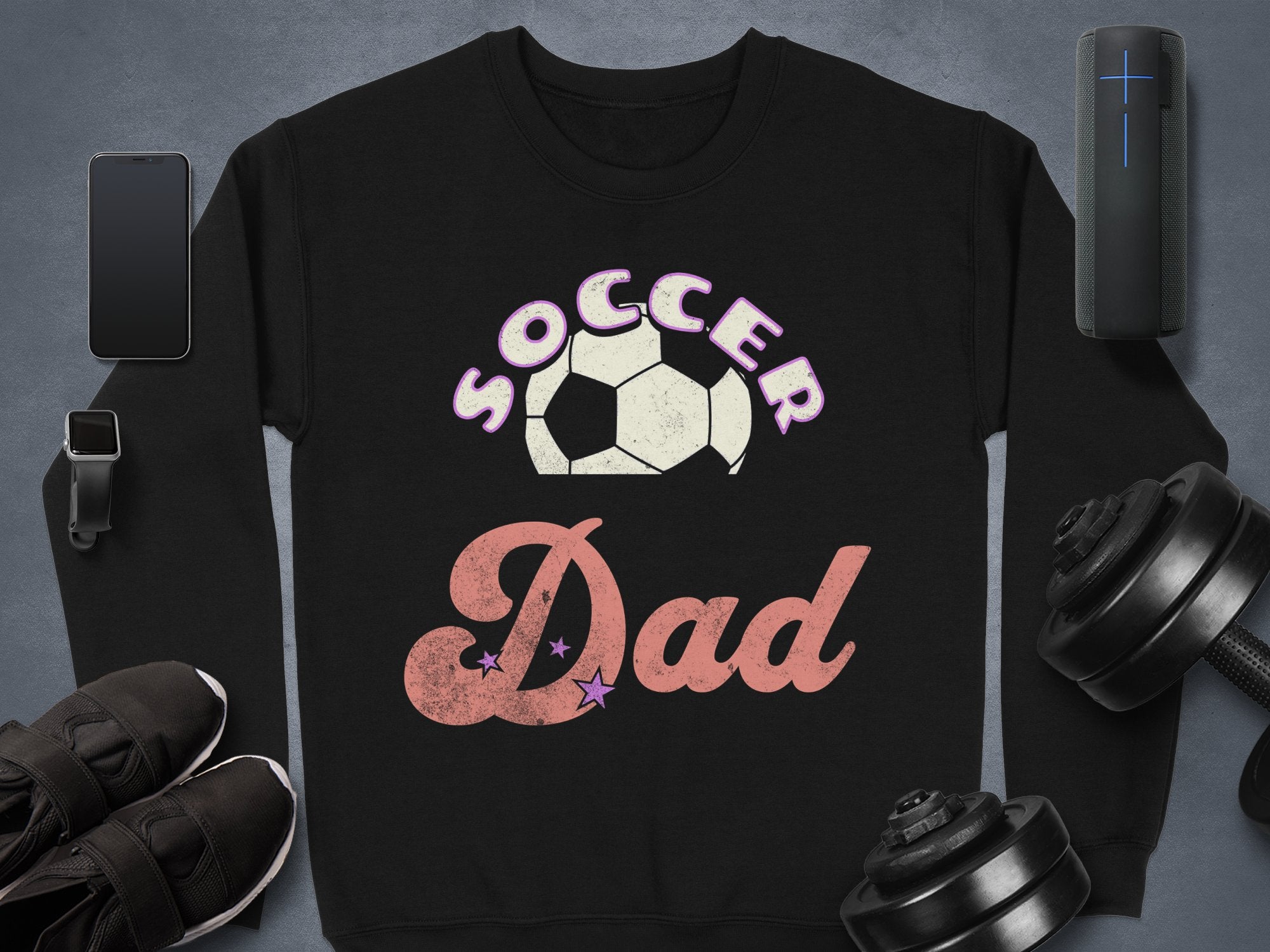 The Soccer Dad sweatshirt by Garment Graphics, showcasing a white and pink soccer ball design, pairs seamlessly with items like smartphones and black sneakers on a grey backdrop. This stylish, cozy piece offers unisex sizing for maximum comfort and versatility.