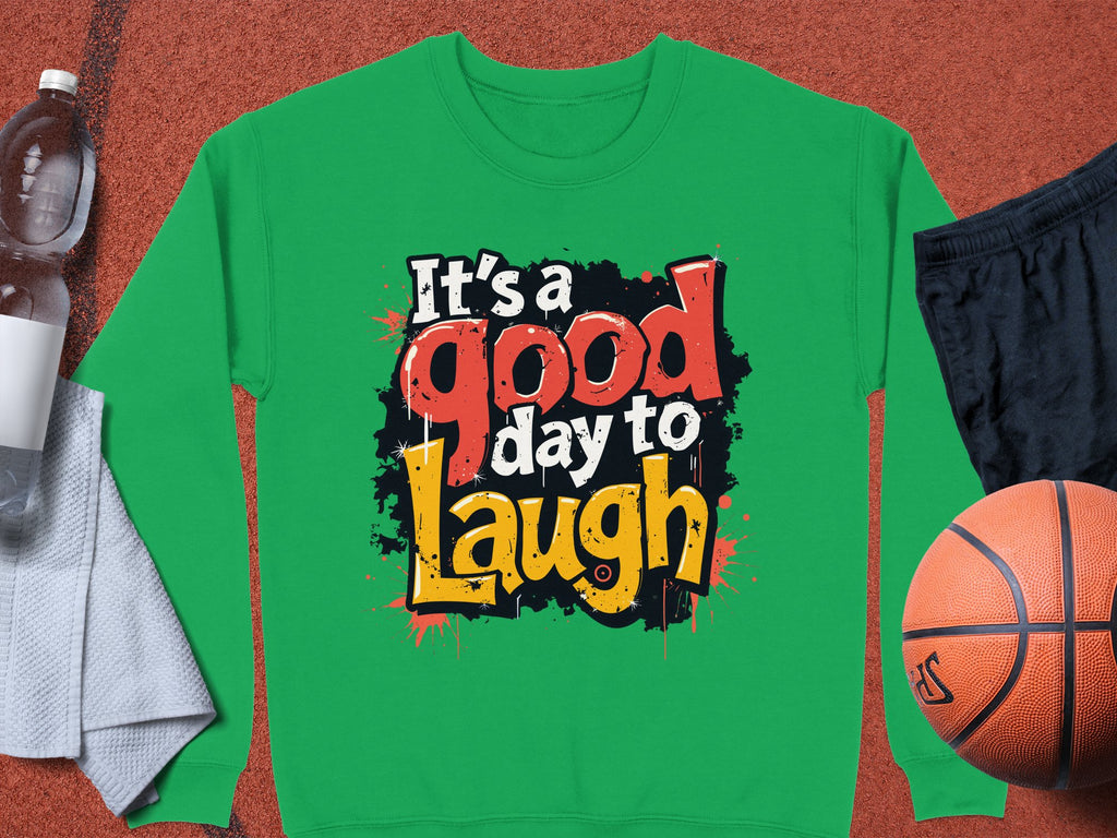 A green sweatshirt from Garment Graphics, featuring the vibrant message Its a Good Day to Laugh, lies next to a basketball, black shorts, a water bottle, and a towel. Unisex sizing ensures its both stylish and comfortable for any playful occasion.