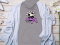 Cozy grey Soccer Mum hoodie by Garment Graphics, featuring pink and white with a soccer ball graphic, rests on wood. Paired with a light blue cardigan, white shoes, an open book, jewelry, and white flowers for a classic ensemble.