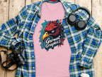 A classic fit pink tee from Garment Graphics features an eagle design and is surrounded by a blue plaid shirt, headphones, a camera, and a coffee cup on a wooden surface.