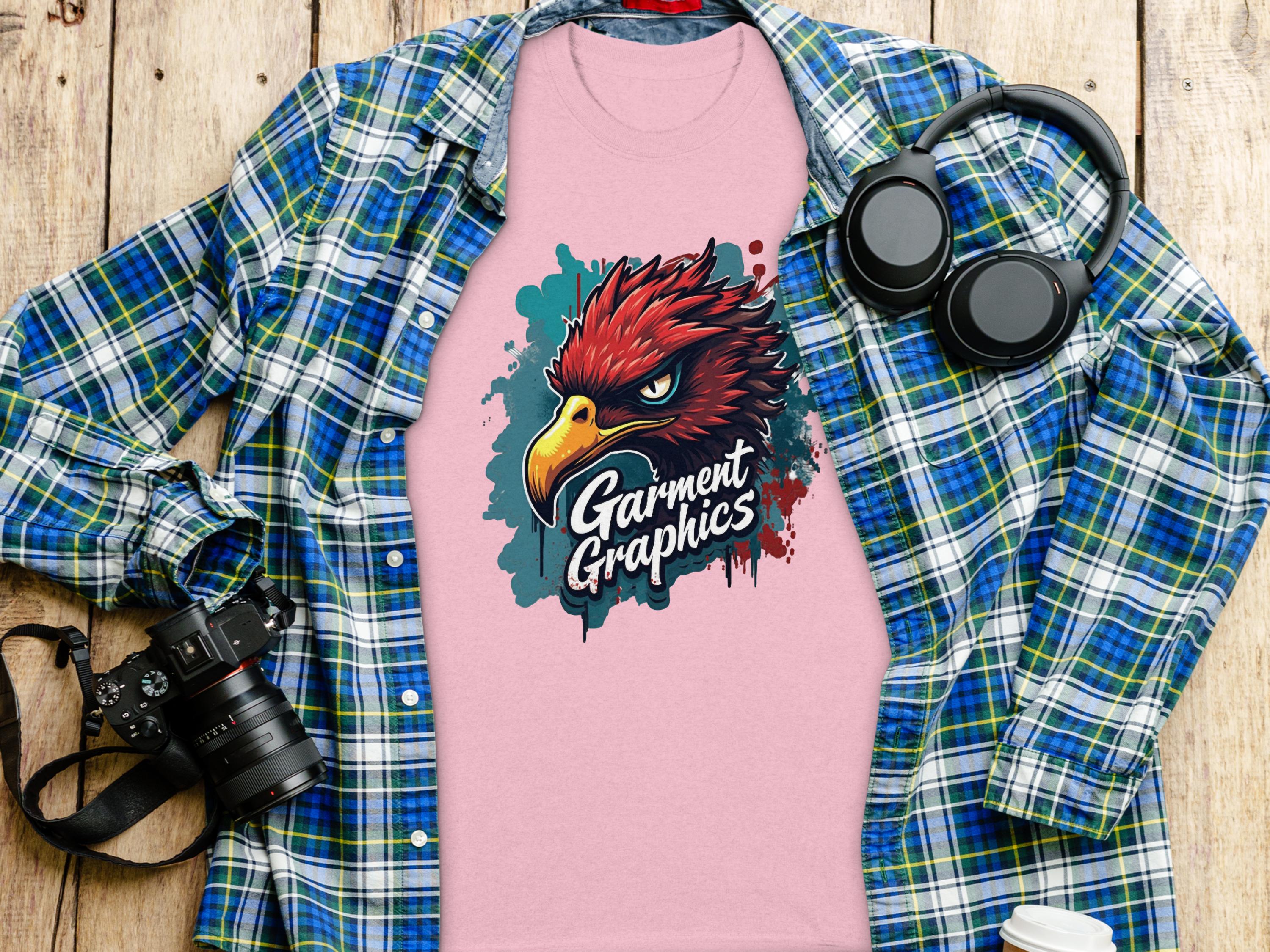 A classic fit pink tee from Garment Graphics features an eagle design and is surrounded by a blue plaid shirt, headphones, a camera, and a coffee cup on a wooden surface.