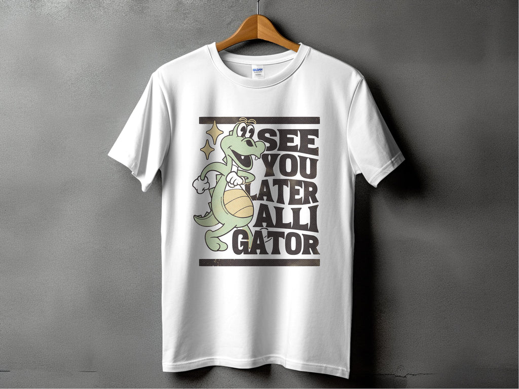 Against a gray background, a white tee by Garment Graphics hangs on a wooden hanger. Ideal for casual outings, it displays a cartoon alligator with a basketball and the phrase SEE YOU LATER ALLIGATOR, encapsulating playful style effortlessly.