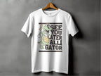 Against a gray background, a white tee by Garment Graphics hangs on a wooden hanger. Ideal for casual outings, it displays a cartoon alligator with a basketball and the phrase SEE YOU LATER ALLIGATOR, encapsulating playful style effortlessly.