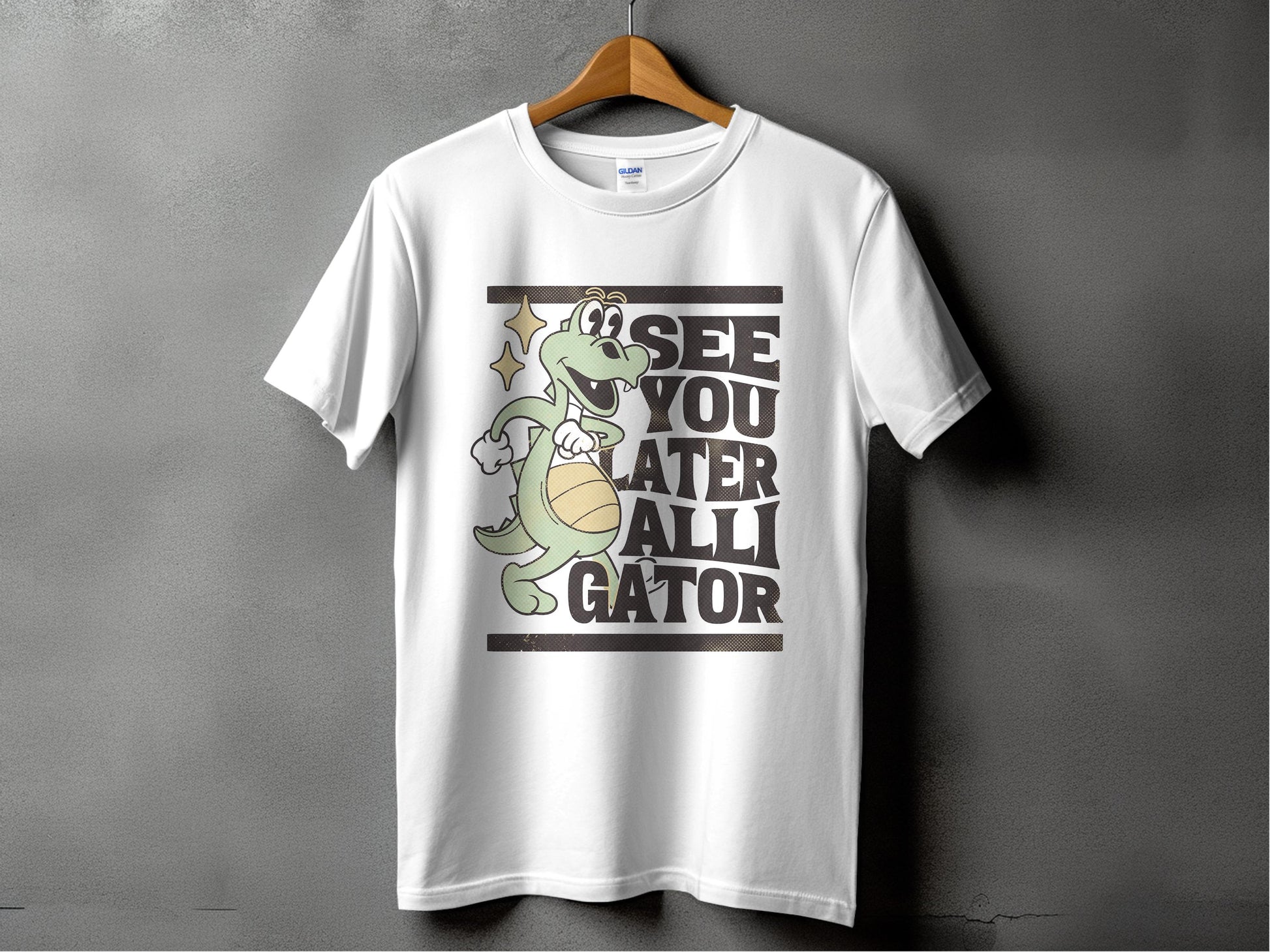Against a gray background, a white tee by Garment Graphics hangs on a wooden hanger. Ideal for casual outings, it displays a cartoon alligator with a basketball and the phrase SEE YOU LATER ALLIGATOR, encapsulating playful style effortlessly.
