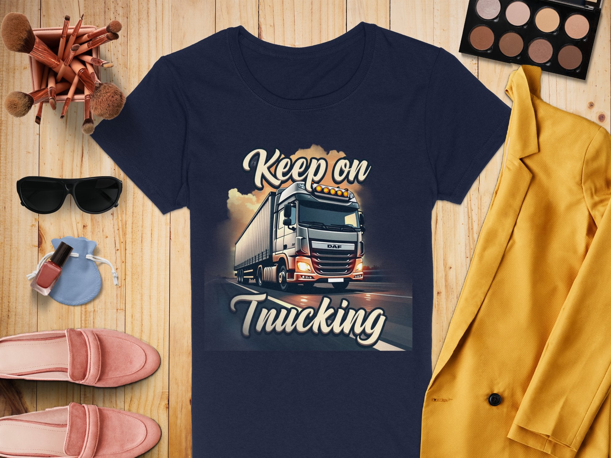A navy Keep on Trucking shirt with a powerful truck design from Garment Graphics is displayed on a wooden surface, surrounded by a yellow blazer, pink loafers, sunglasses, makeup brushes, eyeshadow palette, nail polish, and perfume—ideal for graphic design T-shirt enthusiasts.
