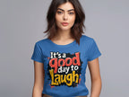 A woman stands against a neutral background, radiating positivity in a semi-fitted blue T-shirt from Garment Graphics, featuring the colorful text Its a good day to laugh.