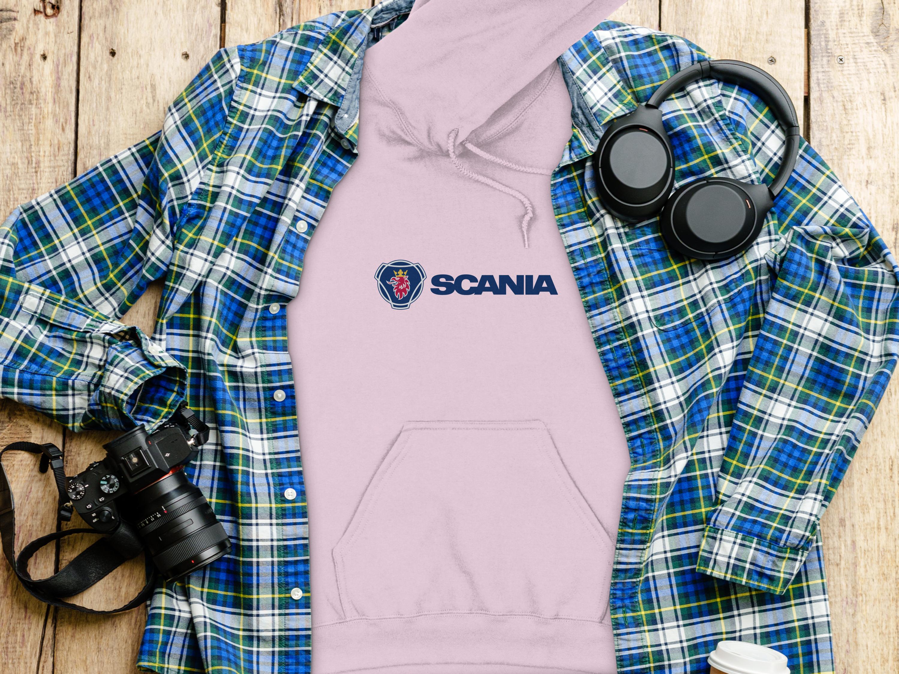 A trendy apparel scene features a pink Garment Graphics hoodie under a blue and green plaid shirt on wood. Nearby are black headphones, a white cup, and a black camera, completing the stylish setup.