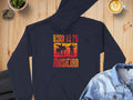 The Garment Graphics black unisex hoodie features Born to Be a Musician text and vibrant red-orange graphics of musicians with instruments. Perfect for music lovers, it is displayed stylishly on wood alongside a denim jacket, coffee, and potted plant.