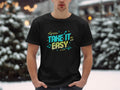 Amidst snowy surroundings, a person wears a Garment Graphics black t-shirt featuring Take It Easy in colorful designs. Snow-covered trees adorn the background, with warm twinkling lights resembling musical notes creating a serene melody.
