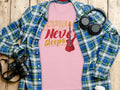 A Garment Graphics 100% cotton pink T-shirt featuring Music Never Sleeps with a guitar design rests on a wooden surface, surrounded by a blue and green plaid shirt, headphones, a camera, and a coffee cup lid.