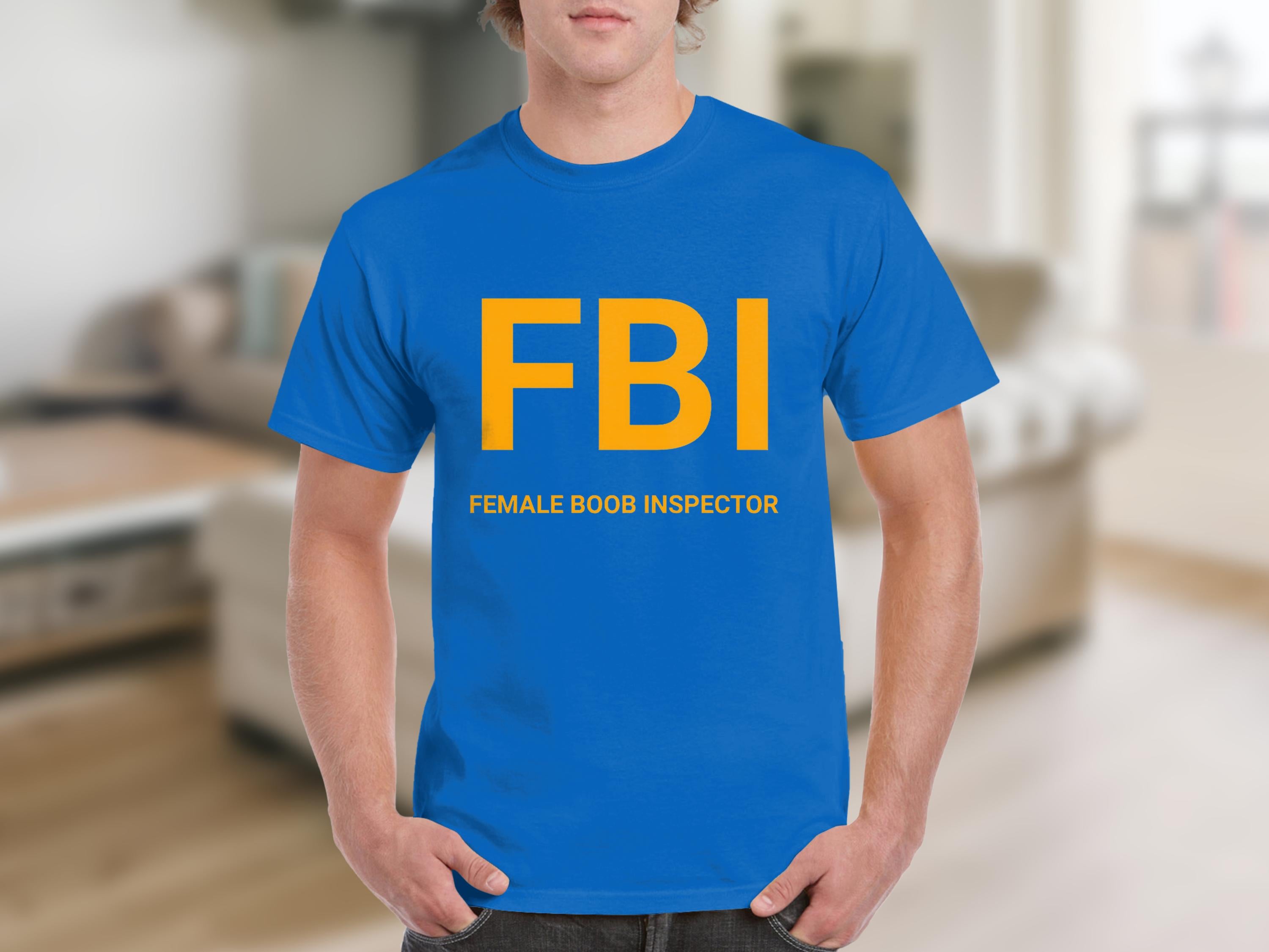 A person wears a Garment Graphics T-shirt featuring the humorous design FBI in large yellow letters, with FEMALE BOOB INSPECTOR captioned below, standing in a blurred indoor setting.