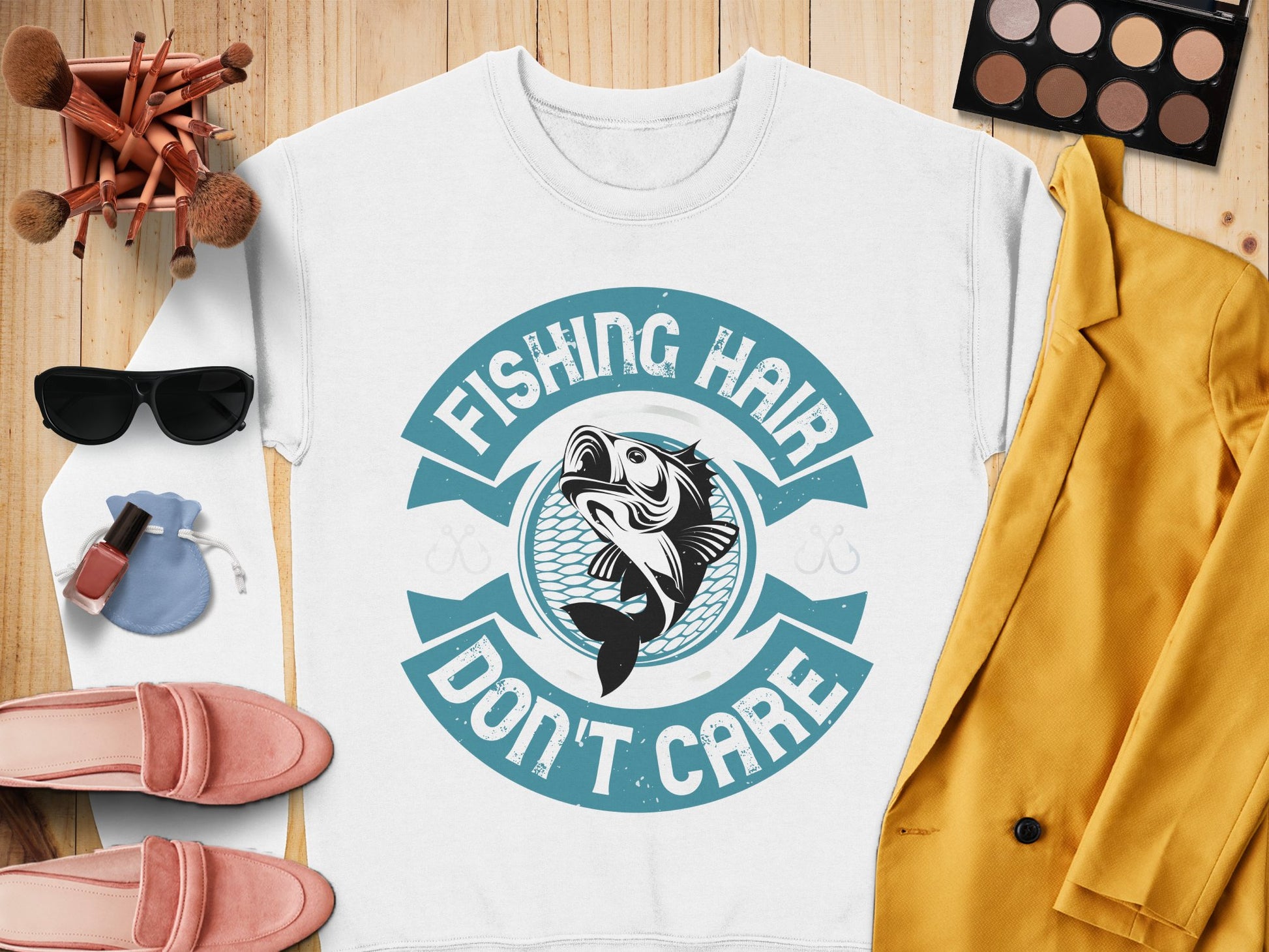 Garment Graphics stylish white sweatshirt with Fishing Hair Dont Care print is perfect for fishing enthusiasts. Presented on a wooden surface, its accompanied by a yellow blazer, pink shoes, makeup brushes, an eyeshadow palette, sunglasses, nail polish, and a blue pouch.