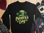 A vibrant black sweatshirt by Garment Graphics features a festive St. Patricks Day design with a green mug, frothy drink, and shamrocks. It sits on wood surrounded by a hat, gloves, plaid shirt, and eyeglasses.