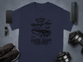 A navy T-shirt by Garment Graphics features a vintage car and Retro Automotive Club text, surrounded by a smartphone, smartwatch, black sneakers, wireless speaker, and dumbbells on a gray surface.