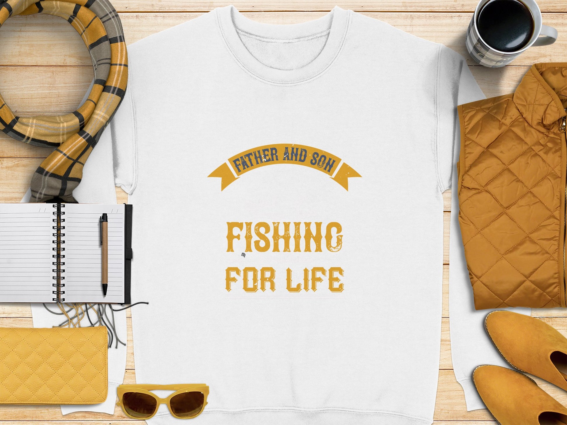 A flat lay from Garment Graphics features a white t-shirt with Father and Son Fishing for Life in yellow. It includes a cozy scarf, notebook, pen, coffee cup, quilted jacket, sunglasses, and wallet in yellow and beige hues—ideal for embodying the father-son adventure spirit.