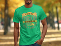 A person outdoors sports a green Garment Graphics T-shirt with the slogan: If hes got golf clubs in his truck or a camper in his driveway, I dont hire him, set against a backdrop of autumn trees lining a pathway, highlighting camping humor.