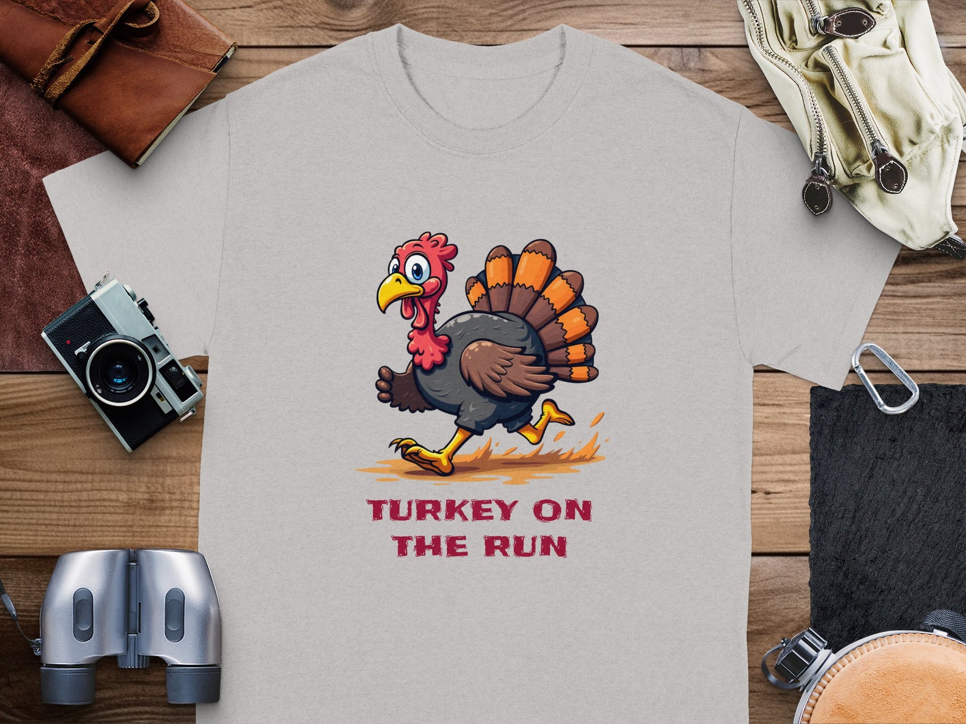 Garment Graphics classic fit gray graphic T-shirt features a funny turkey in red shoes mid-run, with TURKEY ON THE RUN printed below in red. Its displayed on a wooden surface with a camera, binoculars, map, gloves, and pouch.