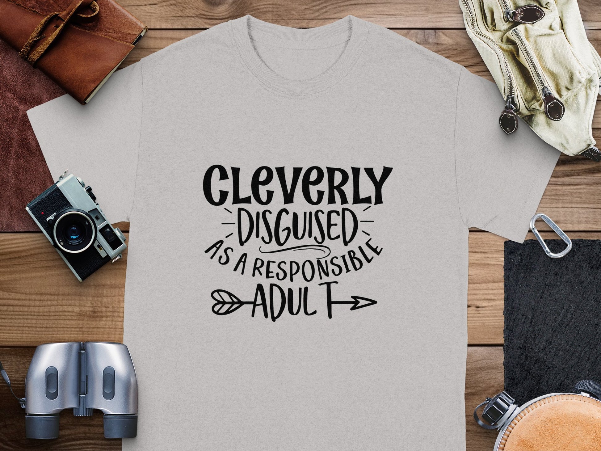 A light gray Cleverly Disguised as a Responsible Adult T-shirt by Garment Graphics, in a classic fit, is ideal for casual outings. Its laid on a wooden surface and surrounded by items like a camera, leather pouch, binoculars, and wristwatch.