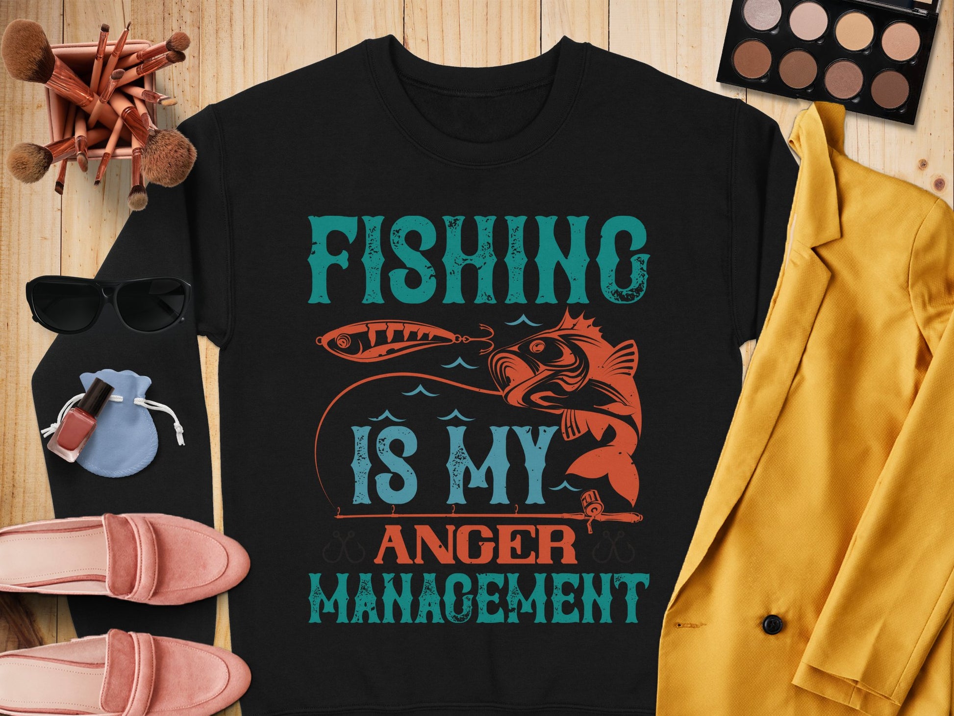 A black shirt from Garment Graphics, featuring Fishing Is My Anger Management with a fish and hook design, lies on a wooden surface. Ideal for fishing fans, its paired with a blazer, makeup palette, brushes, sunglasses, perfume, blue pouch, and pink shoes.