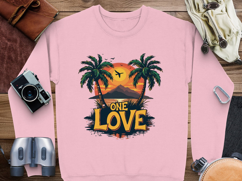 This vibrant pink sweatshirt by Garment Graphics features a One Love Sunset design with silhouetted palm trees against a sunset and bold yellow ONE LOVE letters. Nearby, a camera, binoculars, leather bag, and goggles enhance the adventurous vibe.