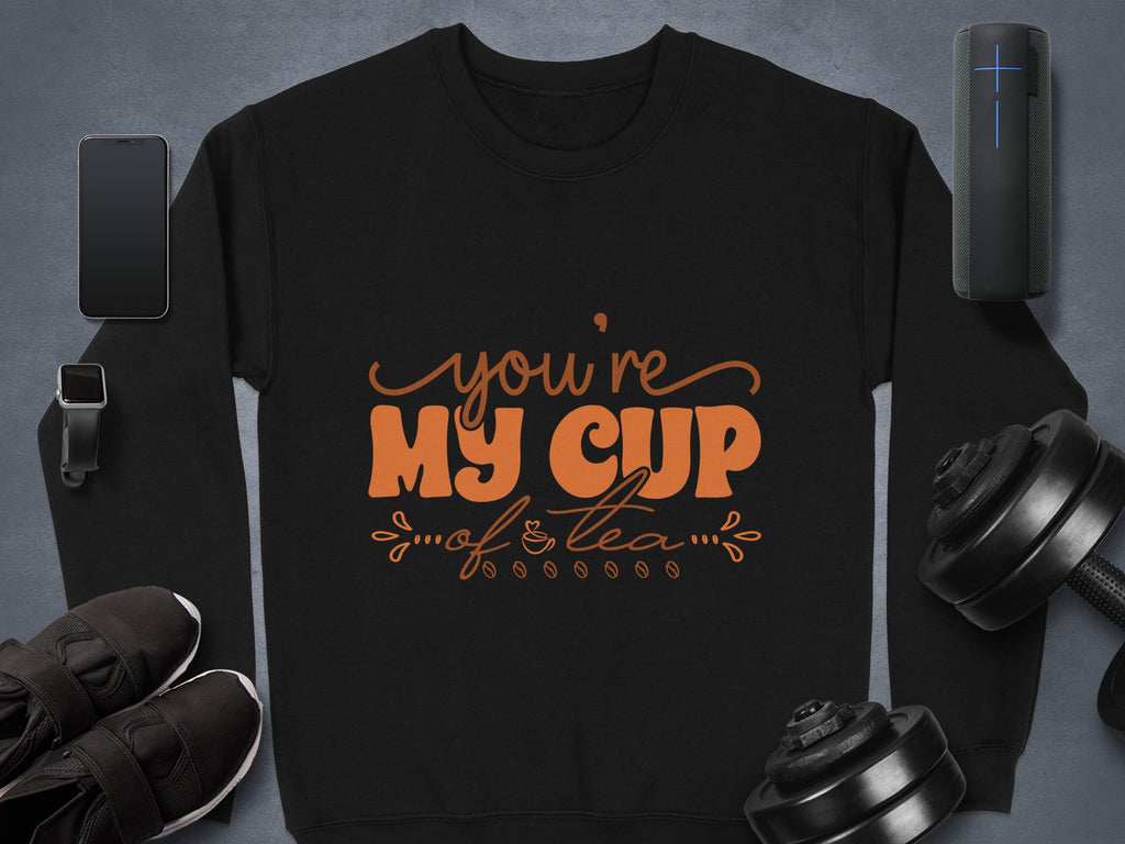 A flat lay image showcases a Garment Graphics cozy black sweatshirt with Youre My Cup of Tea in vibrant orange letters, ideal for tea lovers. Nearby are a smartphone, smartwatch, sneakers, portable speaker, and adjustable dumbbells on a gray surface.