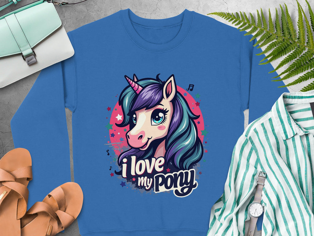 A blue Garment Graphics sweatshirt features a cartoon unicorn with a purple and teal mane, pink horn, and I love my pony text. Its on concrete with sandals, a striped shirt, watch, small bag, and ferns—ideal for unicorn enthusiasts.