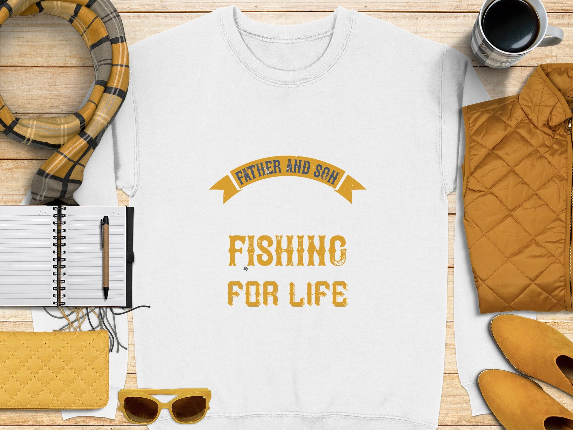 Flat lay showcasing a Garment Graphics white t-shirt with yellow Father and Son Fishing for Life text. Items include a notebook, pen, yellow quilted jacket, graphic sweatshirt, sunglasses, scarf, mug on a wooden background.