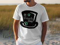A person in a white Garment Graphics T-shirt featuring a large black top hat graphic with shamrocks and St Patricks Day text is in a field of tall grass, enhanced by a subtle four-leaf clover design for added luck.
